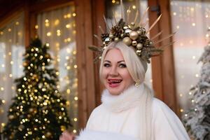 Happy blonde as Snow Maiden in white costume. Wooden house exterior Festive setting. Character with crown creating festive scene. photo