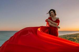 A woman in a red dress is standing on a grassy hillside. The sun is setting in the background, casting a warm glow over the scene. The woman is enjoying the beautiful view and the peaceful atmosphere. photo