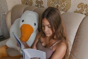A girl is reading a book while sitting on a bed with a stuffed duck. The room has a gold and white color scheme, and there are two lamps in the room. photo