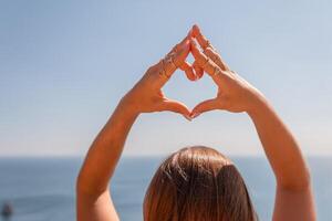 un mujer es participación su manos juntos en un corazón forma, con su brazos extendido hacia el océano. concepto de paz y relajación, como el mujer es disfrutando el belleza de el océano. foto