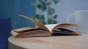 An open book with its pages gently turning rests on a wooden table in the breeze video