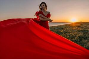 woman red dress standing grassy hillside. The sun is setting in the background, casting a warm glow over the scene. The woman is enjoying the beautiful view and the peaceful atmosphere. photo