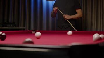 Man in a black vest is seen rubbing the tip of his cue with chalk while standing at a billiard table in a nightclub during a pause in the game. Mid shot. Slow motion video