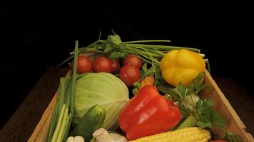 A wooden crate filled with fresh vegetables. The crate contains a variety of colorful produce, including tomatoes, green peppers, yellow peppers, cabbage, cucumbers, scallions, and corn. video