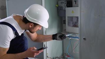 Handsome bearded electrician repairing electrical box. Electrician using screwdriver and a new application on a smartphone video