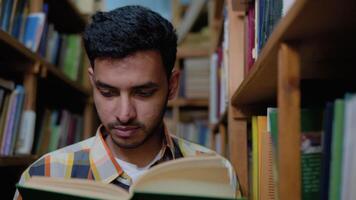 University Library. Talented Indian Boy Standing Next to Bookshelf Reads Book for His Class Assignment and Exam Preparations. Low Angle Portrait video