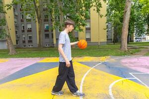 A young man in a grey t-shirt and black pants dribbles a basketball on an outdoor court. The court is painted with yellow, blue, and pink lines. The man is walking towards the right side of the image, with a building complex in the background. photo