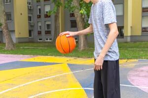 This image shows a young man dribbling a basketball on an outdoor court. He is wearing a gray shirt and black pants. The court is painted in various colors with white lines. There are buildings and green trees in the background. photo