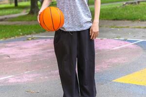 A person, wearing black pants and a gray shirt, is holding a basketball in front of them. They are standing on an outdoor court, and there is a pink and yellow painted area visible in the foreground. photo