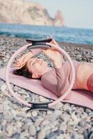 mujer mar pilates deportivo contento medio Envejecido mujer practicando aptitud en playa cerca mar, sonriente activo hembra formación con anillo en yoga estera afuera, disfrutando sano estilo de vida, armonía y meditación foto