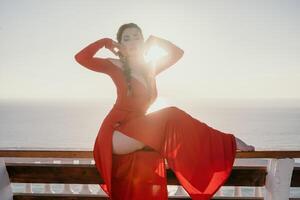 Closeup portrait of sensual young brunette woman in red dress, happily dancing outdoors isolated on blurry sea background with natural bokeh in soft warm sunset backlight. Selective focus. photo