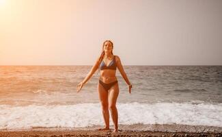 A tourist woman in blue bikini on beach enjoys the turquoise sea during her summer holiday. Rear view. Beach vacation. woman with braids dreadlocks standing with her arms raised enjoying beach ocean. photo