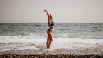 A tourist woman in blue bikini on beach enjoys the turquoise sea during her summer holiday. Rear view. Beach vacation. woman with braids dreadlocks standing with her arms raised enjoying beach ocean. photo