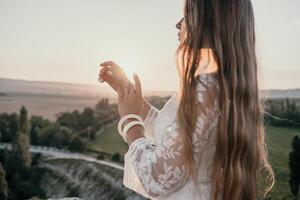 Happy woman in white boho dress on sunset in mountains. Romantic woman with long hair standing with her back on the sunset in nature in summer with open hands. Silhouette. Nature. Sunset. photo