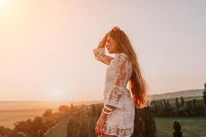 Happy woman in white boho dress on sunset in mountains. Romantic woman with long hair standing with her back on the sunset in nature in summer with open hands. Silhouette. Nature. Sunset. photo