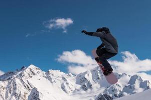 Portrait of a young man snowboarder jumping on a snowboard in sportswear, against the backdrop of high snow-capped mountains and sky photo