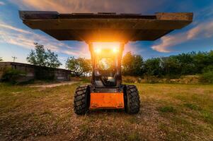 Small tractor loader with a raised bucket at sunset with headlights on. Wide Angle photo