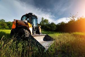 small bulldozer stands in green grass against the backdrop of sun rays in a forest belt at sunset photo