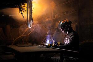 A welder in a protective mask for metal welding and protective gloves in a leather jacket performs welding work in a home garage. Sparks, hot flame from welding. photo
