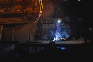A welder in a protective mask for metal welding and protective gloves in a leather jacket performs welding work in a home garage. Sparks, hot flame from welding. photo