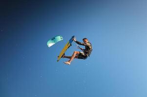 A male kiteboarder in flight over the water does a trick in a clear sky with splashes. Copy space. Kite in the frame. Looking at the camera photo