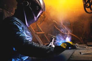A welder in a protective mask for metal welding and protective gloves in a leather jacket performs welding work in a home garage. Sparks, hot flame from welding. photo