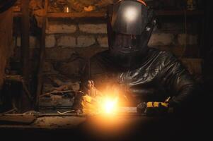 A welder in a protective mask for metal welding and protective gloves in a leather jacket performs welding work in a home garage. Sparks, hot flame from welding. photo