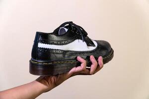 A man's hand holds a black and white brogue wingtip shoe with a rubber outsole made from genuine cowhide. Men's hands holding elegant and shiny two tone shoes on a cream background photo