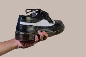 A man's hand holds a black and white brogue wingtip shoe with a rubber outsole made from genuine cowhide. Men's hands holding elegant and shiny two tone shoes on a cream background photo