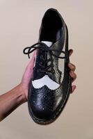 A man's hand holds a black and white brogue wingtip shoe with a rubber outsole made from genuine cowhide. Men's hands holding elegant and shiny two tone shoes on a cream background photo