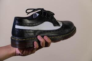 A man's hand holds a black and white brogue wingtip shoe with a rubber outsole made from genuine cowhide. Men's hands holding elegant and shiny two tone shoes on a cream background photo