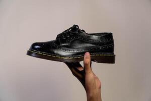 Man's hand holding elegant and shiny full black vintage shoes on a beige background. A jet black brogue wingtip shoe with a rubber outsole is held in a man's hand photo