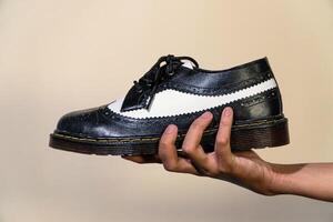 A man's hand holds a black and white brogue wingtip shoe with a rubber outsole made from genuine cowhide. Men's hands holding elegant and shiny two tone shoes on a cream background photo