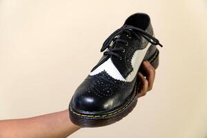 A man's hand holds a black and white brogue wingtip shoe with a rubber outsole made from genuine cowhide. Men's hands holding elegant and shiny two tone shoes on a cream background photo