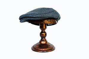 Detail of a scally cap or flat hat made of wool herringbone mounted on a bronze mannequin head on a white background. Catalog photo of a classic dark gray hat on an elegant bronze mannequin