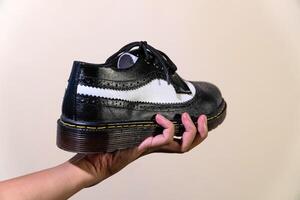 A man's hand holds a black and white brogue wingtip shoe with a rubber outsole made from genuine cowhide. Men's hands holding elegant and shiny two tone shoes on a cream background photo