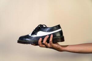 A man's hand holds a black and white brogue wingtip shoe with a rubber outsole made from genuine cowhide. Men's hands holding elegant and shiny two tone shoes on a cream background photo