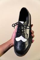 A man's hand holds a black and white brogue wingtip shoe with a rubber outsole made from genuine cowhide. Men's hands holding elegant and shiny two tone shoes on a cream background photo