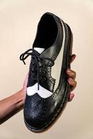 A man's hand holds a black and white brogue wingtip shoe with a rubber outsole made from genuine cowhide. Men's hands holding elegant and shiny two tone shoes on a cream background photo