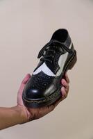 A man's hand holds a black and white brogue wingtip shoe with a rubber outsole made from genuine cowhide. Men's hands holding elegant and shiny two tone shoes on a cream background photo