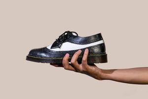 A man's hand holds a black and white brogue wingtip shoe with a rubber outsole made from genuine cowhide. Men's hands holding elegant and shiny two tone shoes on a cream background photo