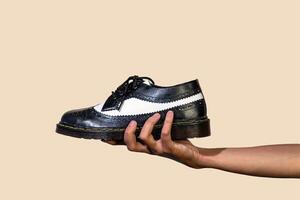 A man's hand holds a black and white brogue wingtip shoe with a rubber outsole made from genuine cowhide. Men's hands holding elegant and shiny two tone shoes on a cream background photo