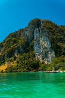 Large rock cliff covered in green trees with the sea in front of it on a sunny day. The view of large rock cliffs and vast ocean is a tourist icon for tropical islands in Krabi, Thailand photo