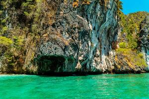 Large rock cliff covered in green trees with the sea in front of it on a sunny day. The view of large rock cliffs and vast ocean is a tourist icon for tropical islands in Krabi, Thailand photo