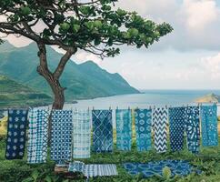 Beautifully patterned blue fabrics hanging on a clothesline under a tree, set against a peaceful mountain backdrop near the sea. photo