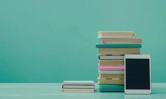 Stack of books on a light gray table with a pastel turquoise background with a tablet beside photo