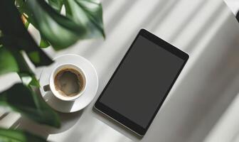 Tablet on a light blue table with a cup of tea on the right, shadow from window, top view photo