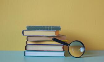 Stack of books on a light gray table with a pastel butter yellow background, with a magnifying glass photo