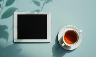 Tablet on a light blue table with a cup of tea on the right, shadow from window, top view photo