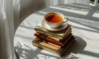 Stack of books with a cup of tea on a light gray table, soft shadows from window photo